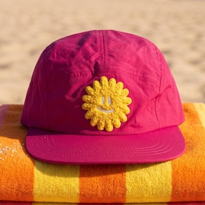 May include: A stack of four striped beach towels in orange, yellow, and pink, topped with a magenta cap. The cap features a yellow smiling flower applique. The items are arranged on a sandy beach, suggesting a summer theme.