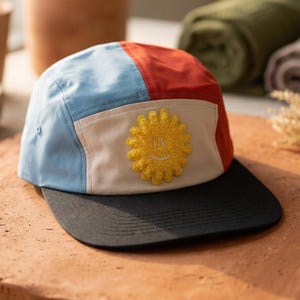 May include: A colorful five-panel cap with a black brim. The cap features panels of light blue, red, and beige, with a yellow embroidered sun design on the front. The hat is designed for casual wear.
