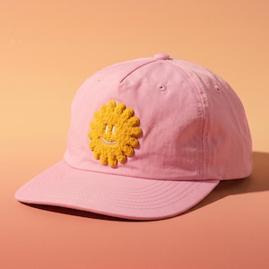 May include: A pink baseball cap with a yellow, smiling flower patch. The cap has a curved brim and a button on top. The background is a gradient of peach and orange.