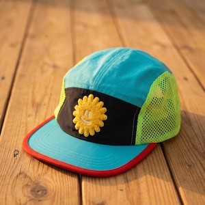 May include: A colorful cap with a turquoise crown, a black band, and a yellow mesh side panel. The cap features a yellow smiling sun patch and a red brim. The design is playful and vibrant.