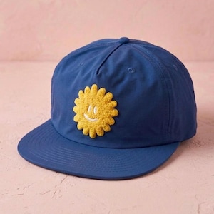 Puede incluir: Una gorra de béisbol azul marino con un parche central de sol amarillo texturizado que presenta una cara sonriente bordada en blanco. La gorra tiene una corona estructurada de cinco paneles, una visera plana y costuras azules a juego.
