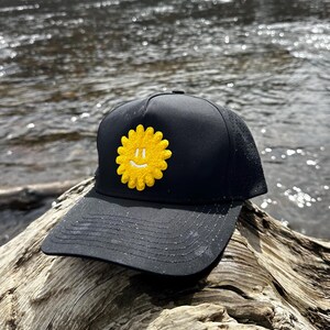 May include: Black baseball cap with a yellow smiling sun patch. The cap has a curved brim and a mesh back for breathability. The sun patch is a bright yellow color with a smiling face.