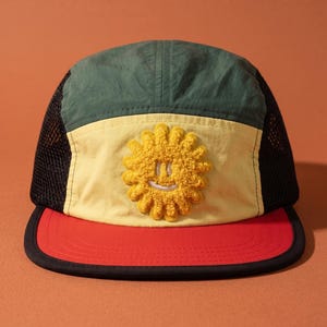 May include: A multi-colored cap with a mesh back and a curved brim. The cap features a red brim, a yellow front panel with a yellow smiling sun design, and a green top panel. The mesh back is black.
