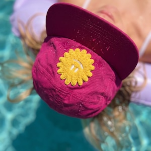 Puede incluir: Gorra de béisbol magenta con un diseño de flor amarilla. La gorra la lleva alguien en una piscina. La flor tiene un centro blanco. La visera de la gorra es curva. El fondo es una piscina.