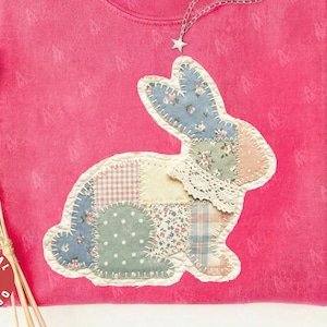 Peut inclure: Un sweat-shirt rose Comfort Colors avec un appliqué de lapin en patchwork. Le lapin est composé de différents tissus à motifs, dont des motifs floraux et vichy. Un collier étoile argenté et un chapeau marron sont également visibles.