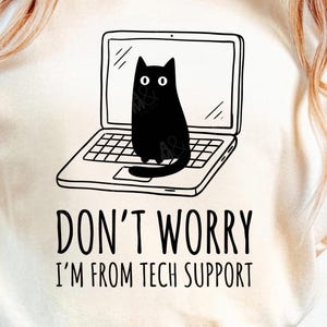 May include: Cream-colored t-shirt featuring a black graphic of a cat sitting on a laptop. The text "DON'T WORRY I'M FROM TECH SUPPORT" is printed below the laptop. The shirt has short sleeves and a crew neck.
