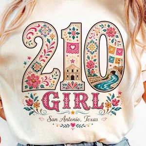 Könnte beinhalten: Cremefarbenes T-Shirt mit einem farbenfrohen Grafikdesign. Das Design zeigt die Zahlen "210" mit floralen und dekorativen Elementen, das Wort "GIRL" und den Text "San Antonio, Texas".