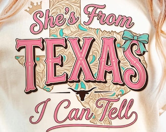 May include: A cream-coloured t-shirt featuring the text "She's From Texas I Can Tell" in pink and brown. The design includes a map of Texas with turquoise accents and a decorative bow. A crown is above the text.