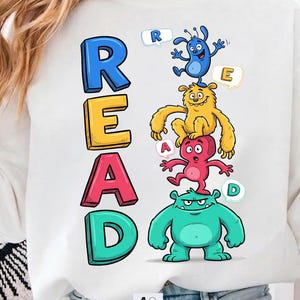 May include: White sweatshirt featuring the word "READ" in colorful, stacked letters. The design includes cartoon monsters, each holding a letter. The monsters are blue, yellow, red, and green. The sweatshirt has a crew neck.