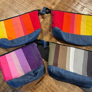 May include: Four colourful zippered pouches with a denim base. The pouches feature vertical stripes in shades of red, orange, yellow, pink, purple, and neutral tones. The pouches are arranged on a wooden surface.