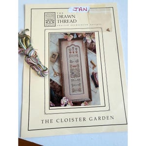 May include: A needlework pattern booklet titled The Cloister Garden by The Drawn Thread, featuring a framed sampler design with floral and geometric motifs. The booklet includes a skein of variegated embroidery thread and a small plastic bag of beads.