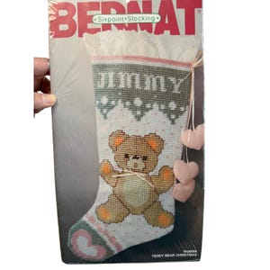 May include: A Bernat Sixpoint Stocking needlepoint kit in original packaging. The stocking features a white background with a brown teddy bear graphic, a green patterned cuff, and the name Jimmy. Three small pink heart charms are attached.