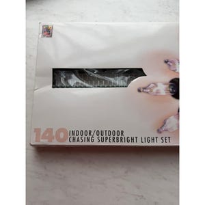 May include: A white box with the text "140 Indoor/Outdoor Chasing Superbright Light Set." The box contains a set of lights. The lights are visible through a rectangular opening in the box.