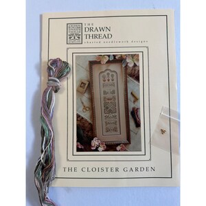 May include: A needlework pattern booklet titled The Drawn Thread The Cloister Garden featuring a sampler design. Included is a skein of variegated embroidery floss in shades of green, pink, and brown, and a small clear bag containing a tiny metal charm.