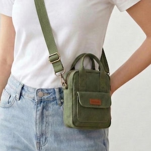 May include: Olive green crossbody bag with a small front pocket and top handle. The bag has a brown leather patch and a matching adjustable strap. The bag is worn over the shoulder. The brand logo "SewLuna" is visible on the upper right.