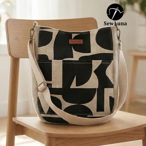 May include: A beige and black patterned shoulder bag with a cream strap and a small "handmade" tag. The bag is on a wooden chair. The bag features a geometric design.
