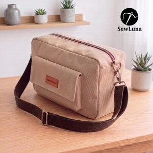May include: A rectangular beige crossbody bag with a dark brown adjustable strap and a dark brown zipper. The bag features a front pocket with a small brown rectangular label that reads handmade in black text. The bag has a structured design.