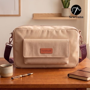 Op de afbeelding: Een rechthoekige beige schoudertas met een voorvak met klep, voorzien van een bruin lederachtig label met de tekst "handmade". De tas heeft donkerpaarse verstelbare zijbanden met metalen clips en een ritssluiting aan de bovenkant.