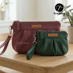May include: Two handmade fabric pouches in burgundy and green, with a pleated design. Each pouch has a wrist strap and a small "handmade" label. The larger pouch is burgundy, and the smaller one is green. The brand name "SewLuna" is visible.