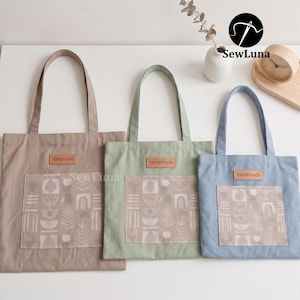 May include: Three canvas tote bags in beige, sage green, and light blue. Each bag has a matching pocket with a geometric pattern and a leather tag that says "handmade". The bags are displayed on a white surface.