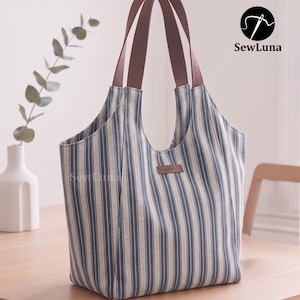 Puede incluir: Un bolso tote a rayas azules y blancas con asas marrones. El bolso tiene forma rectangular con una abertura ancha y una pequeña etiqueta de cuero que dice "handmade". El bolso está sobre una mesa de madera.