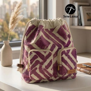 Peut inclure: Un sac à dos à cordon de serrage avec un motif géométrique violet et crème. Le sac a deux poches avant avec des fermetures éclair dorées et une petite étiquette "handmade". Le haut est de couleur crème.