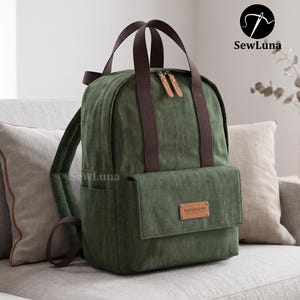 May include: A forest green backpack featuring dark brown webbing straps, two top handles, and a front pocket with a rectangular leather-like patch embossed with the word handmade. The main compartment has dual zippers with light brown pull tabs.