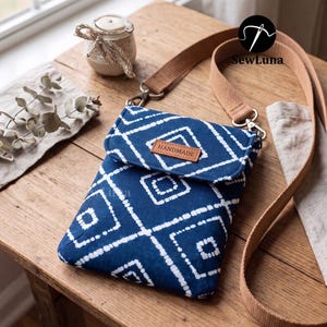 May include: A blue and white patterned crossbody bag with a brown strap and a flap closure. The bag features a geometric design and a small leather tag that reads "HANDMADE". The bag is on a wooden table.