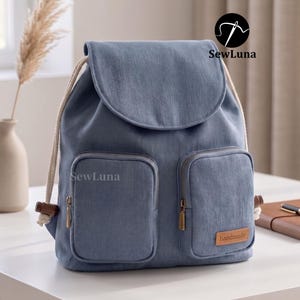 May include: A blue backpack with two front pockets and a flap closure. The bag has a drawstring closure and wooden toggles. The brand name "SewLuna" is visible on the bag. A small leather tag reads "handmade".