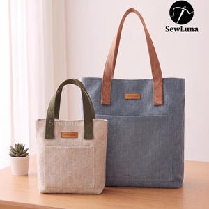 May include: Two tote bags, one small and one large, sit on a wooden table. The larger bag is blue with brown handles and a front pocket. The smaller bag is beige with green handles and a front pocket. Both bags have a "handmade" tag.