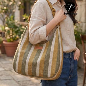 May include: A striped tote bag with a shoulder strap. The bag features vertical stripes in shades of olive green, mustard yellow, and cream. A small, rectangular tag reads "handmade". The bag is carried by a person wearing a light-coloured shirt and blue jeans.