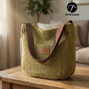 May include: A green canvas tote bag with a brown strap and a small brown leather patch that says "handmade". The bag is sitting on a wooden table. The bag has a rectangular pocket on the front.