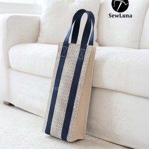 Tall fabric wine bottle carrier with a grey geometric pattern and navy straps, standing on a wooden table next to a wine bottle tied with a red ribbon. Modern kitchen background. Represents a digital sewing pattern for a reusable wine tote bag.