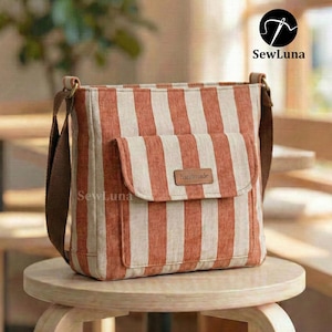 May include: A crossbody bag with orange and cream stripes. It has a front pocket with a flap and a brown strap. A small leather tag reads "handmade". The bag is displayed on a small wooden stool.