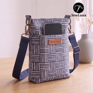 May include: A vertical crossbody bag featuring a blue and white geometric basketweave pattern. The bag includes a front pocket containing a smartphone and a brown leather-like tag labeled Hand Made. It has a dark blue adjustable strap with metal hardware.