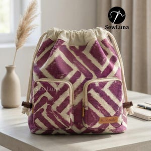 May include: A drawstring backpack with a purple and white geometric pattern. The bag has two front pockets with zippers and a small leather tag that reads "handmade". The bag is sitting on a light gray surface.