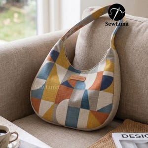 May include: A handmade, geometric-patterned shoulder bag in shades of yellow, blue, orange, and gray. The bag features a curved shape, a long strap, and a small "handmade" tag. The bag is resting on a beige sofa.