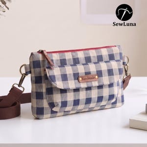 Puede incluir: Un bolso bandolera rectangular con un patrón de cuadros vichy en azul y crema. El bolso incluye un bolsillo frontal con solapa, una etiqueta de cuero sintético marrón con la palabra handmade, cremallera roja y una correa de tela ajustable.