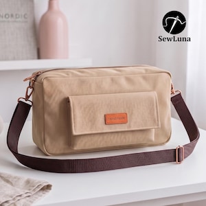 May include: A beige rectangular crossbody bag with a dark brown adjustable strap and copper-toned hardware. The bag features a front flap pocket with an orange rectangular label that reads "handmade" in black text and a top zipper closure.