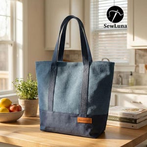 Op de afbeelding: Een denim tote bag met een tweekleurig ontwerp, met een lichtblauwe bovenkant en een donkerblauwe basis. De tas heeft stevige handvatten en een klein "handmade" label. De tas staat op een houten aanrecht.