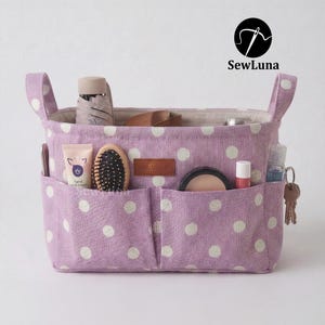 Purple polka dot handmade fabric purse organizer bag filled with daily essentials like a hairbrush, hand cream, and keys. A PDF sewing pattern project for a bag insert or desk storage bin displayed on a clean white background.