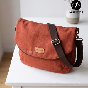Puede incluir: Un bolso tipo mensajero de color óxido con acabado de tela texturizada, que cuenta con una correa ajustable de lona marrón oscuro, herrajes de latón envejecido y una etiqueta rectangular con la palabra handmade grabada en el centro.