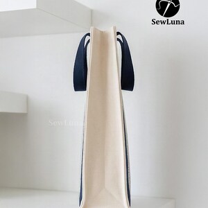 May include: A beige tote bag with navy blue handles and trim. The bag is rectangular with a flat bottom and is displayed against a white background. The SewLuna logo is visible in the upper right corner.