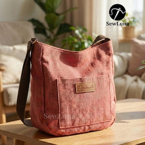 May include: A dusty rose-colored shoulder bag with a brown strap and a front pocket. The bag has a rectangular leather patch that reads "handmade". The bag is sitting on a wooden table.