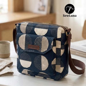 May include: A navy blue crossbody bag with a geometric pattern of white and beige semi-circles and squares. The bag features a front pocket, a brown leather strap, and a zip closure. The word "handmade" is on a small leather tag.