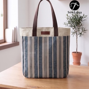 May include: A tote bag featuring a blue and beige vertical striped pattern on the lower section and a solid beige upper band. It has two dark brown fabric handles and a small brown leather-like tag labeled handmade on the front center.