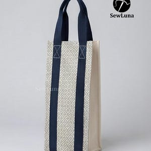 Tall fabric wine bottle carrier with a grey geometric pattern and navy straps, standing on a wooden table next to a wine bottle tied with a red ribbon. Modern kitchen background. Represents a digital sewing pattern for a reusable wine tote bag.