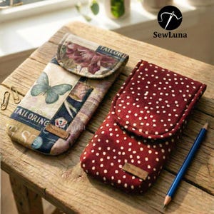 Op de afbeelding: Twee handgemaakte stoffen hoesjes met klepsluitingen. Eén hoesje heeft een vlinder- en bloemenontwerp, de andere is rood met witte stippen. Een blauw potlood ligt naast het rode hoesje. De hoesjes liggen op een houten oppervlak.