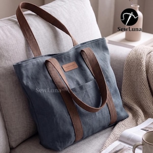 May include: A blue tote bag with brown straps and a front pocket rests on a couch. The bag has a small, rectangular tag that reads "handmade". The bag is made of a textured fabric and is branded with the SewLuna logo.