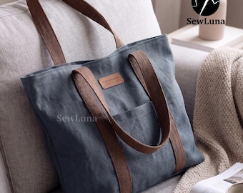 May include: A blue tote bag with brown straps and a front pocket rests on a couch. The bag has a small, rectangular tag that reads "handmade". The bag is made of a textured fabric and is branded with the SewLuna logo.